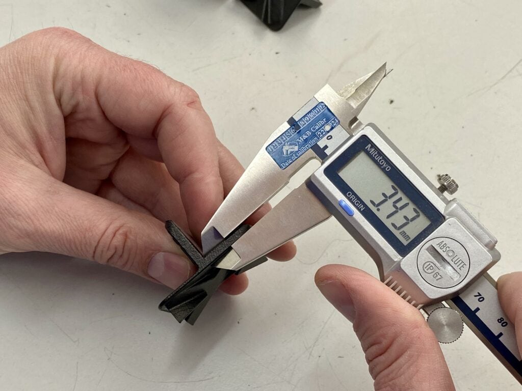 A person using a digital caliper to measure the thickness of a black 3D-printed part, with the caliper