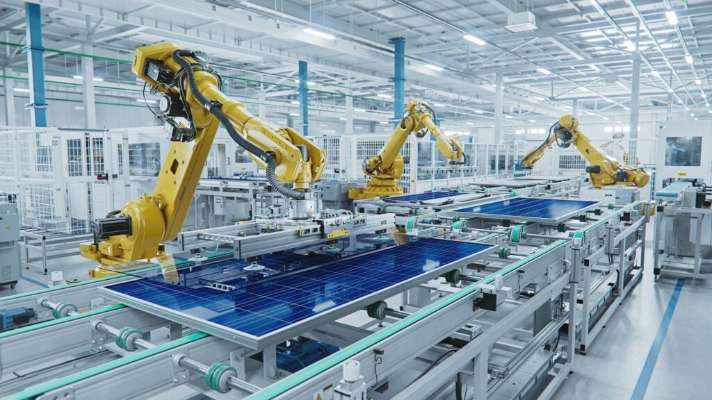 A modern industrial setting where yellow robotic arms work on an assembly line, handling large solar panels. The bright, spacious facility is equipped with advanced automation machinery, likely for manufacturing or assembling solar energy products. 