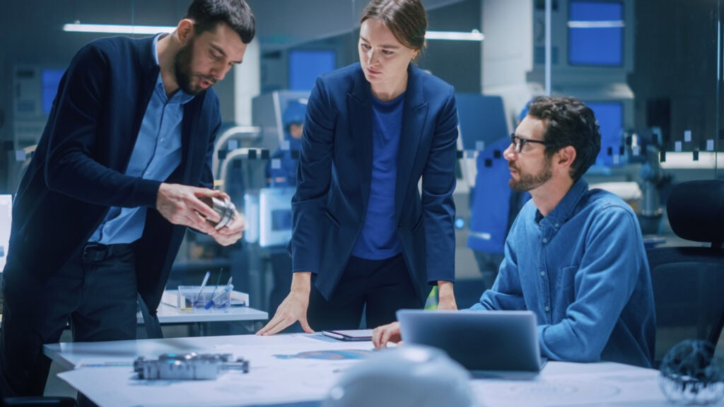 Three engineers collaborate in a modern R&D facility, with one man holding a machined metal part