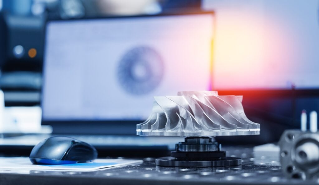 Close-up of a complex 5-axis CNC machined aluminum impeller on a workbench, with its CAD wireframe visible on a laptop screen in the background.